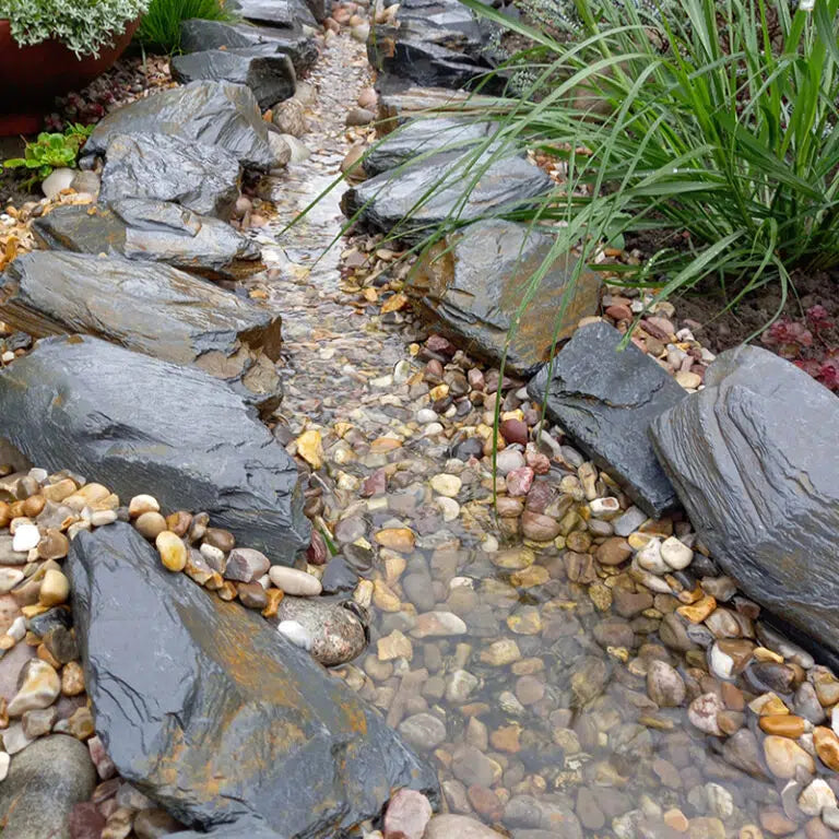 Slate stones edging a shallow pebble-lined garden stream beside planting