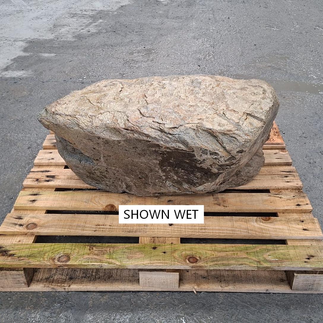 A large wet decorative rock with a rough, textured surface sits on a wooden pallet outdoors.