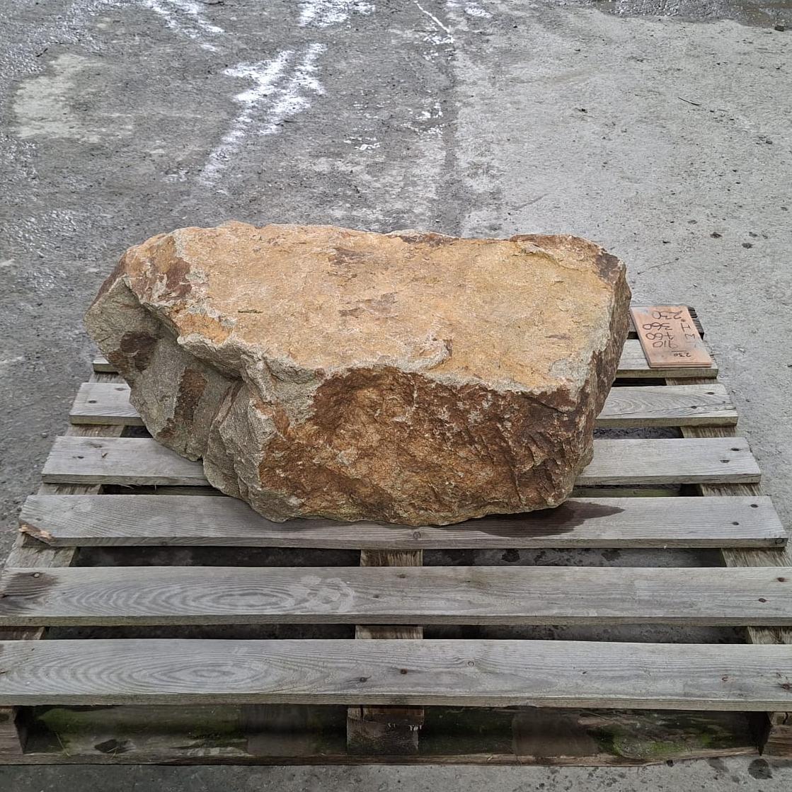 Back view of a large natural stone boulder with rough tan, brown, and grey weathered surfaces, resting on a wooden pallet outdoors.