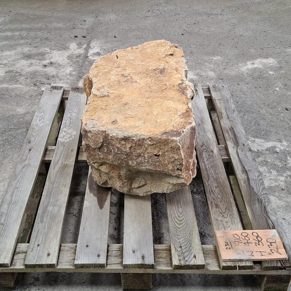 Side view of a large natural stone boulder with rough tan, brown, and grey weathered surfaces. The flat top of the stone can be seen clearly.