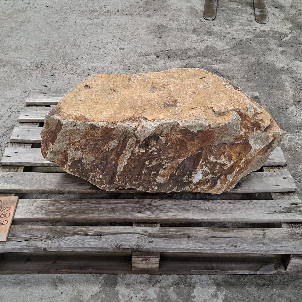 A large natural stone boulder with rough tan, brown, and grey weathered surfaces, resting on a wooden pallet outdoors.