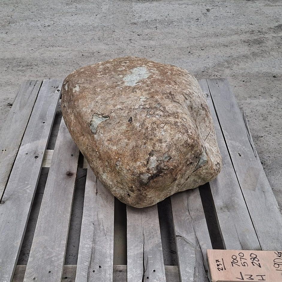 A rough natural stone boulder with a mottled tan, brown, and grey surface sits on a wooden pallet outdoors.