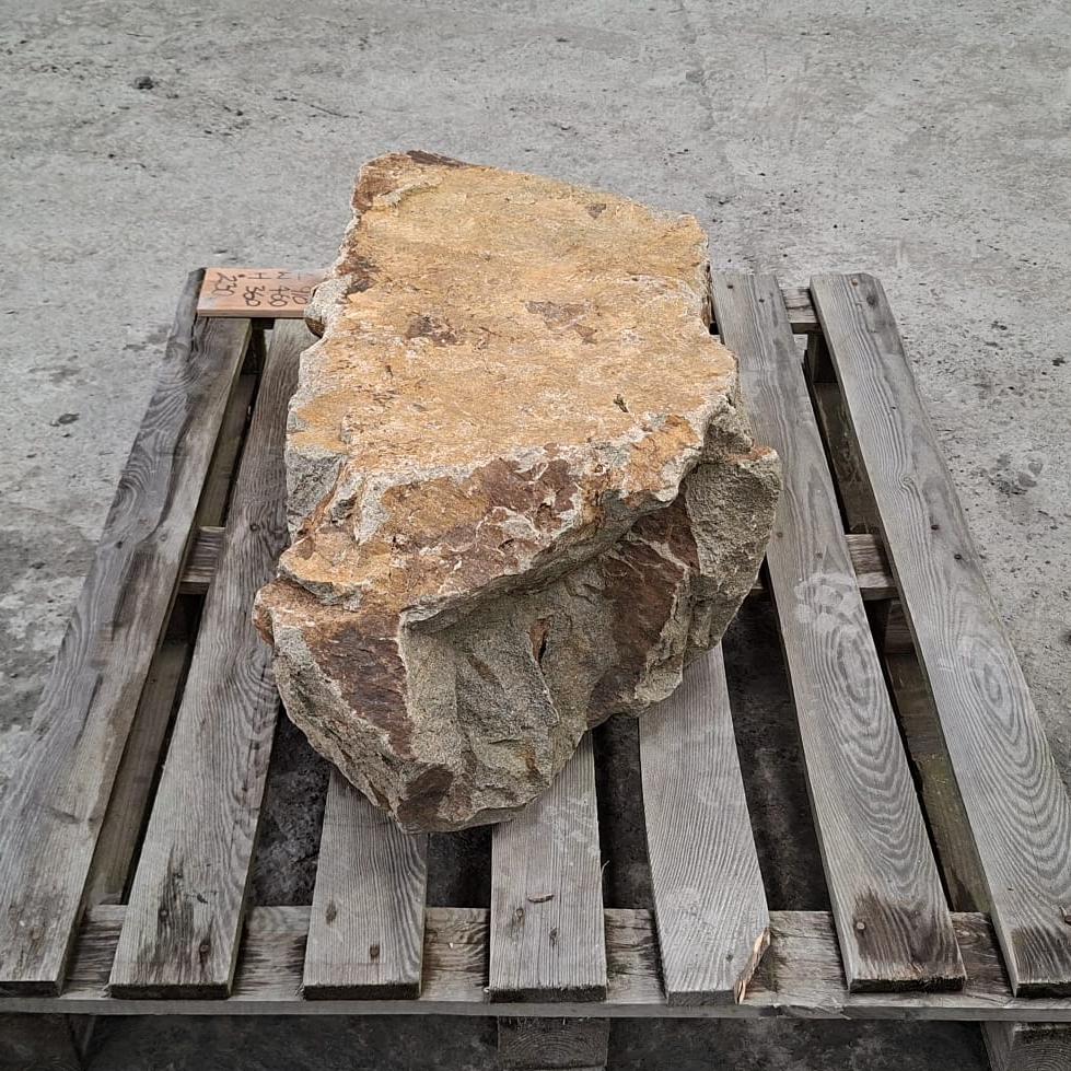 Side view of a large natural stone boulder with rough tan, brown, and grey weathered surfaces, resting on a wooden pallet outdoors.