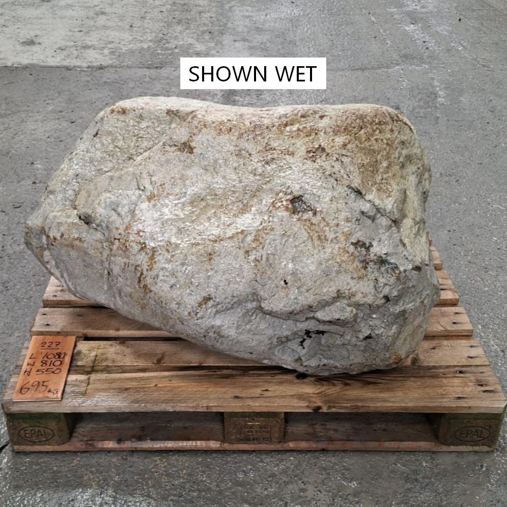 A large wet stone or boulder with a rough, light gray to off-white surface and brownish patches, displayed on a wooden pallet.